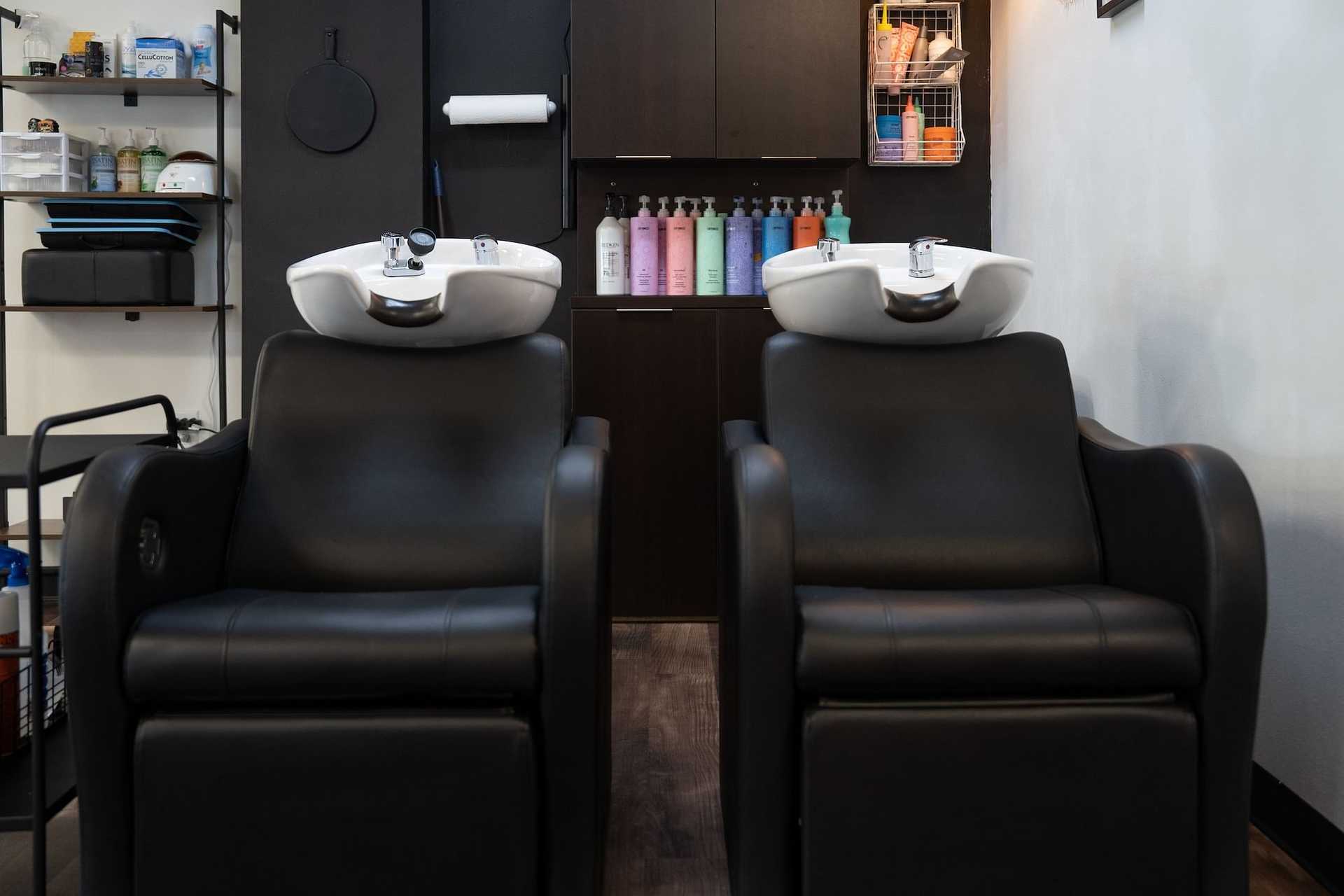 Two black salon chairs in front of hair washing sinks, with hair products displayed behind them.