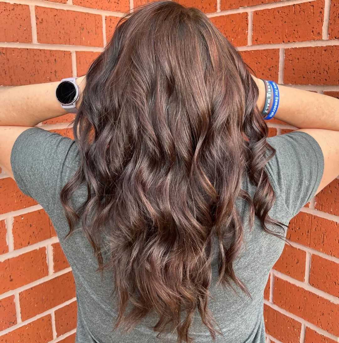 Back view of a person with wavy brown hair against a brick wall, wearing a smartwatch and bracelet.