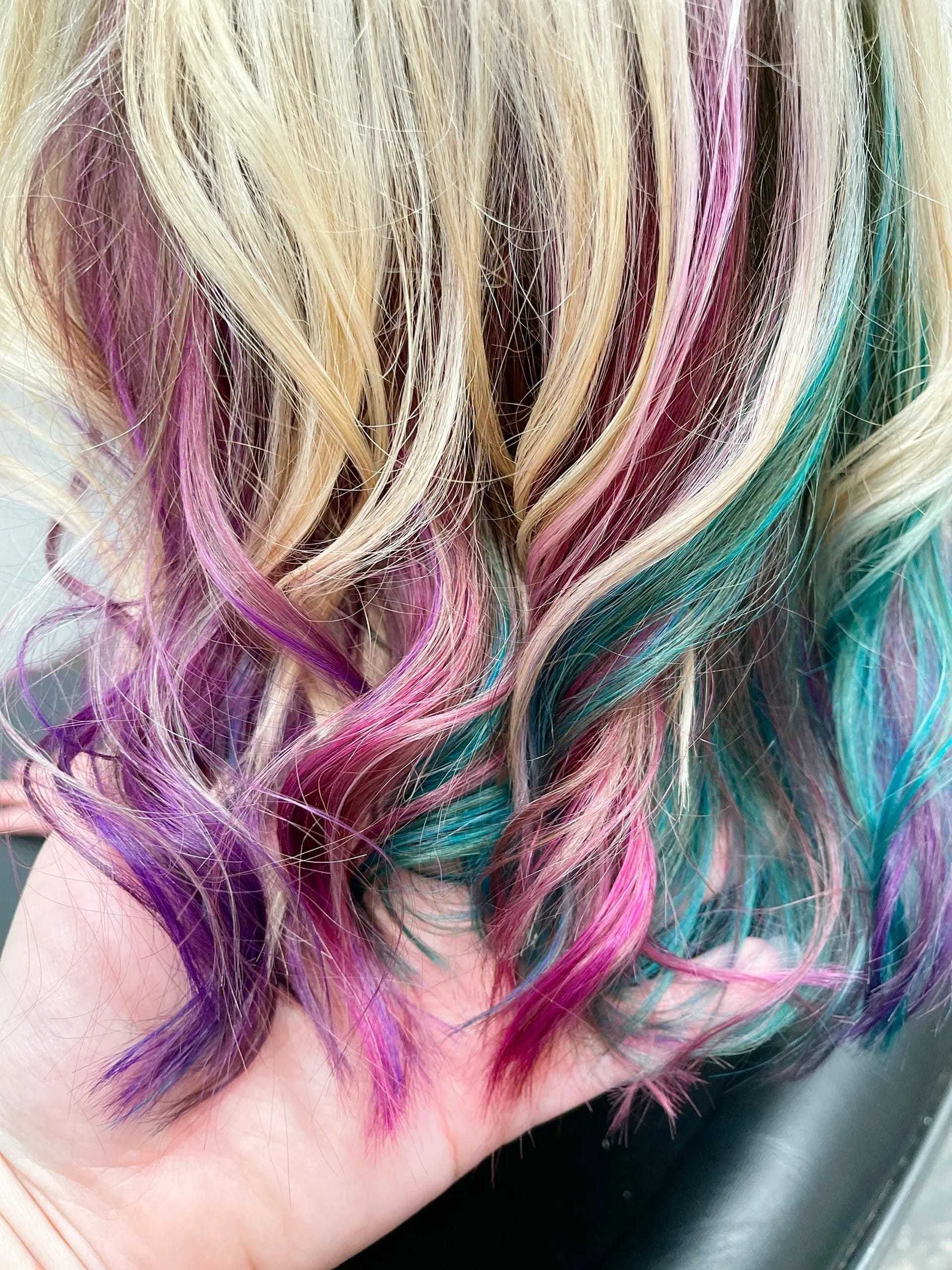 Blonde hair with vibrant purple, pink, blue, and green highlights, styled in loose waves.