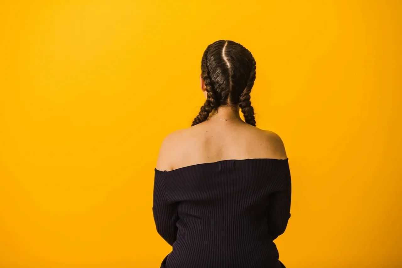 Person with braided hair facing a bright yellow wall, wearing an off-shoulder black top.