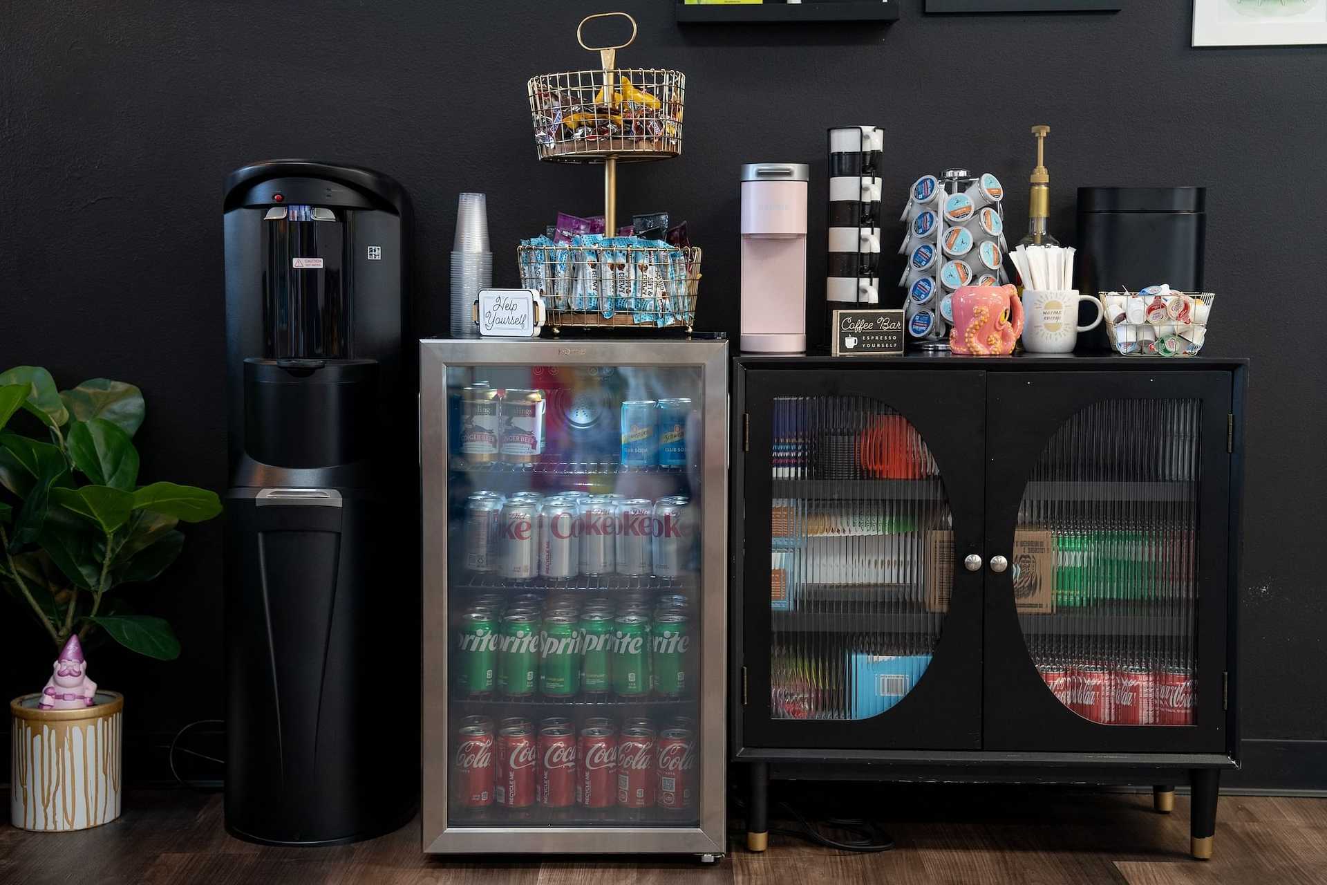 Office breakroom with stocked beverage fridge, coffee maker, water dispenser, and snack baskets.