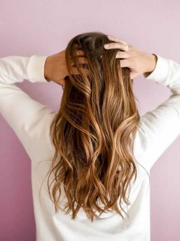 Woman with long, wavy hair facing away, hands on head against a light purple background.