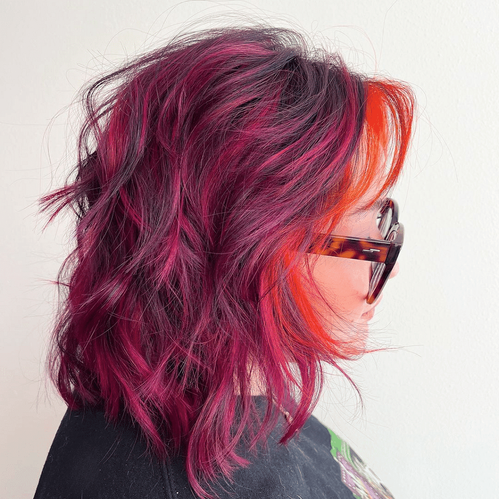 Person with short, wavy, pink and orange hair wearing sunglasses, viewed from the side.